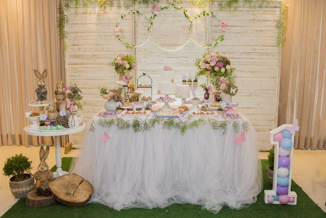 Luxurious table of sweets and birthday cake decorated with flower arrangements and delicate wild animals with light wood panel in the background