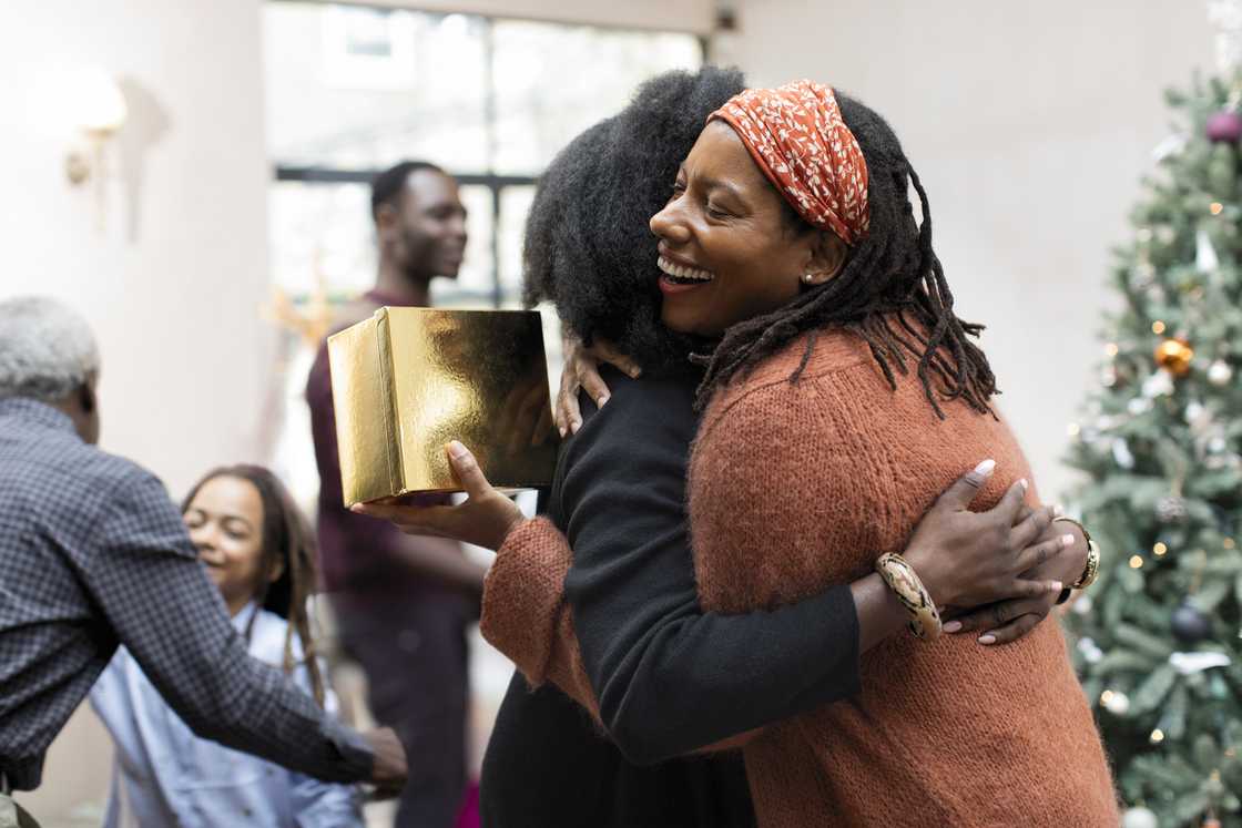 Two people embrace joyfully indoors near a decorated Christmas tree, one holding a gold-wrapped gift.