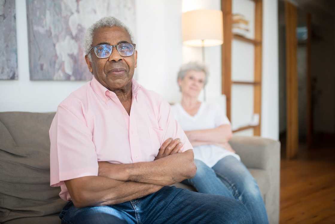 An older man sitting with folded arms while a woman sits behind him on a sofa. An older man sitting with folded arms while a woman sits behind him on a sofa.