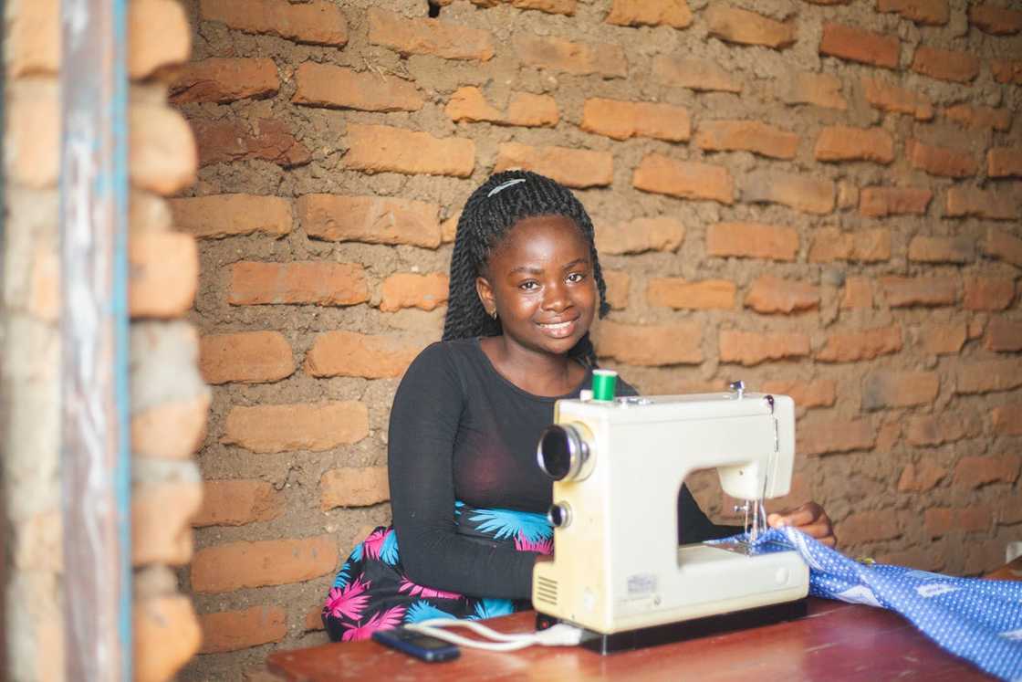 A young woman smiles while seated at a sewing machine indoors. A young woman smiles while seated at a sewing machine indoors.