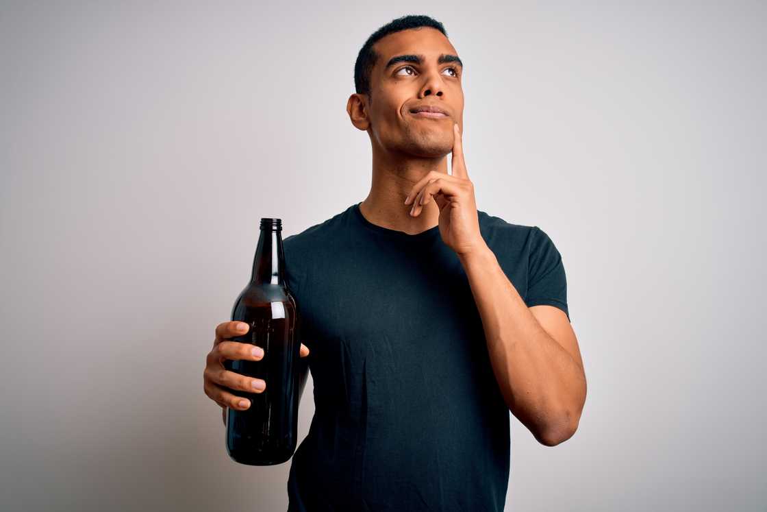 Young African American man drinking a bottle of beer over a white background, looking very confused
