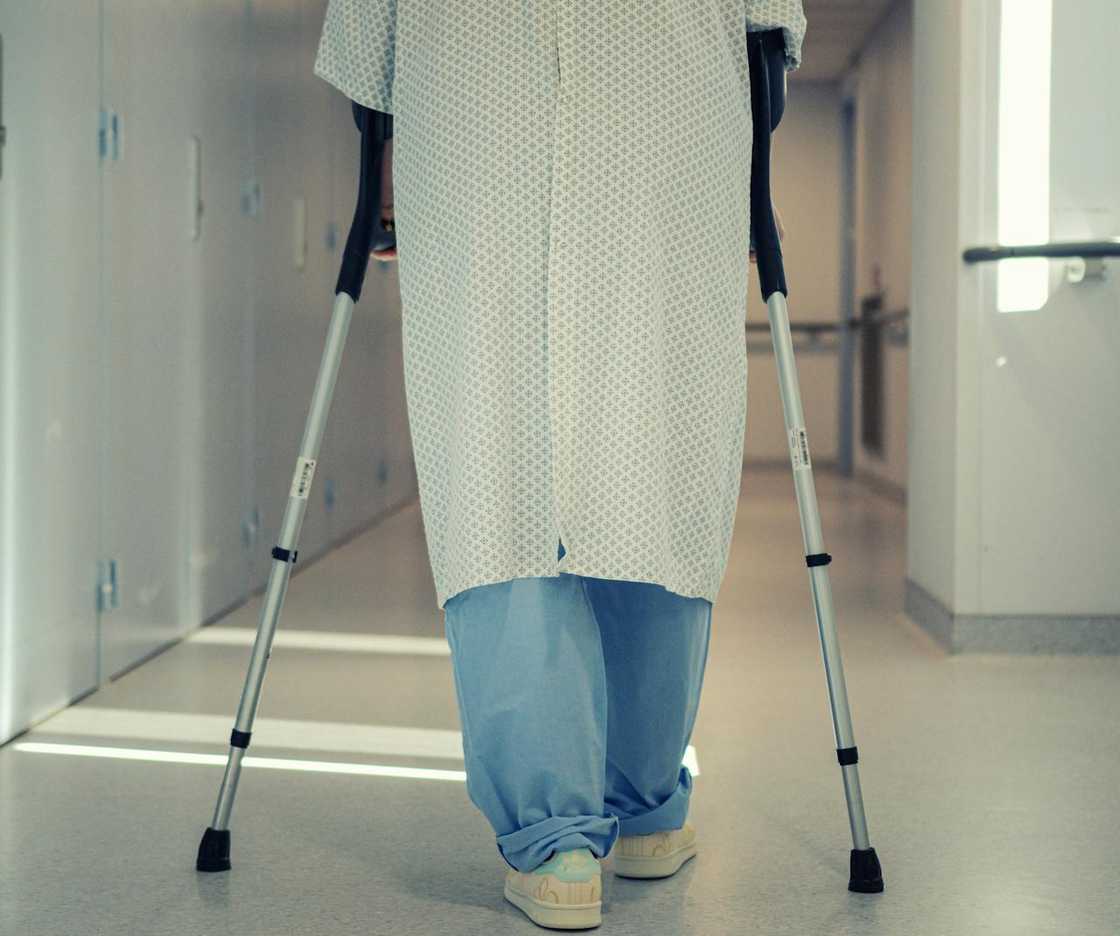 A patient in a hospital gown walks down a corridor using crutches. A patient in a hospital gown walks down a corridor using crutches.