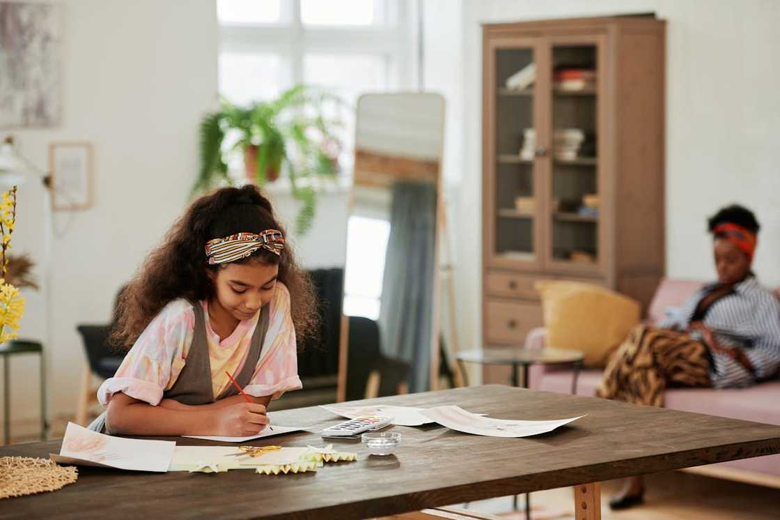 A girl draws at a table while an adult sits in the background.
