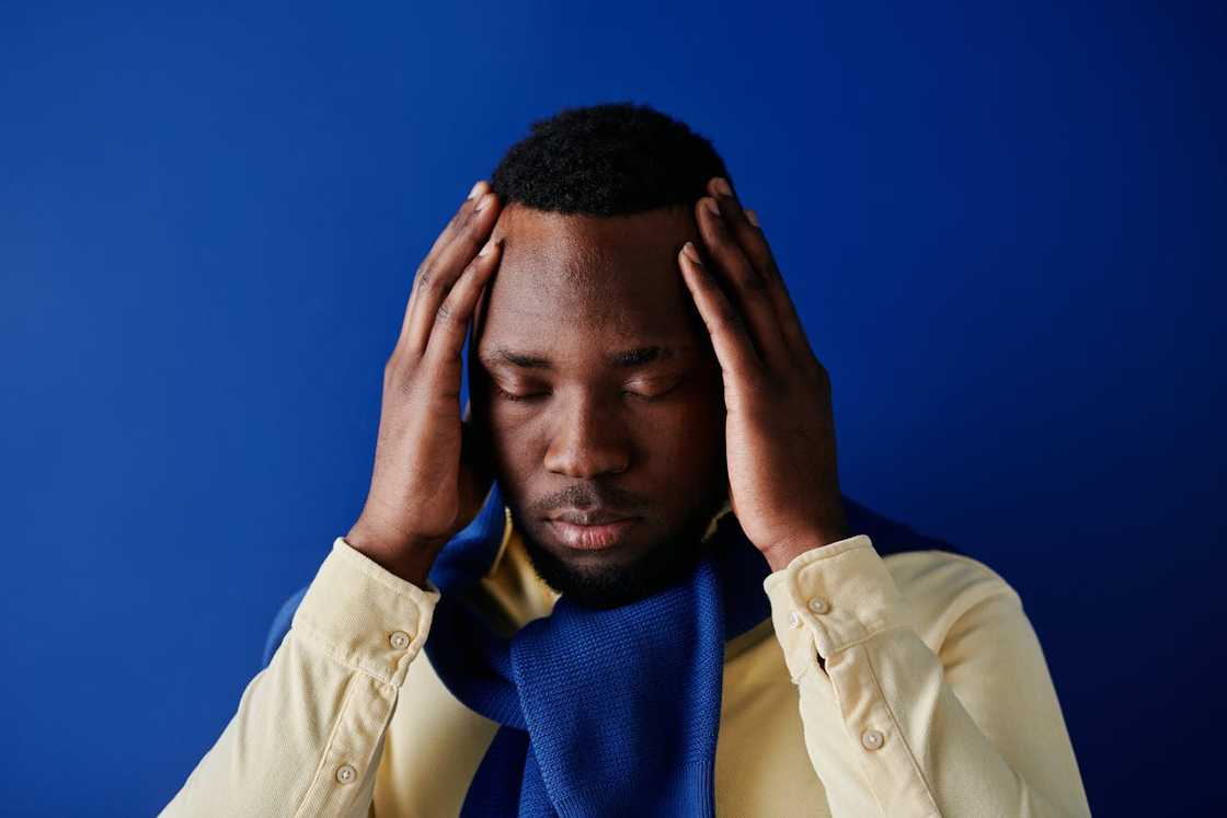 A man holds his head with both hands, eyes closed, against a blue background. A man holds his head with both hands, eyes closed, against a blue background.