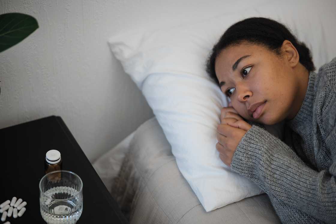 A sick woman lying on bed near bedside table with pills and glass of water A sick woman lying on bed near bedside table with pills and glass of water