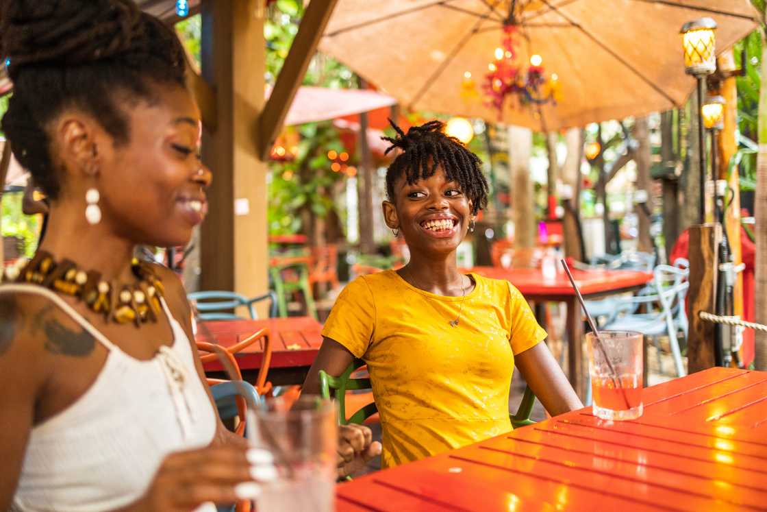 Two people sit at a red outdoor café table, smiling and enjoying drinks under decorative lights. Two people sit at a red outdoor café table, smiling and enjoying drinks under decorative lights.