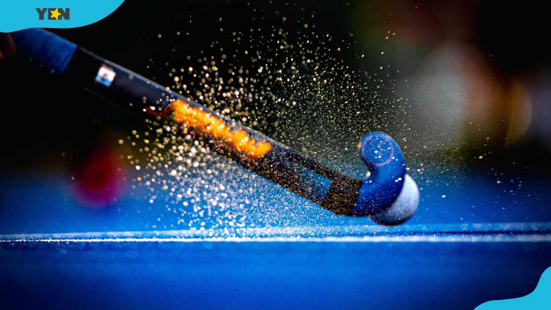 A hockey stick hits a ball on a wet field lit by the sun.