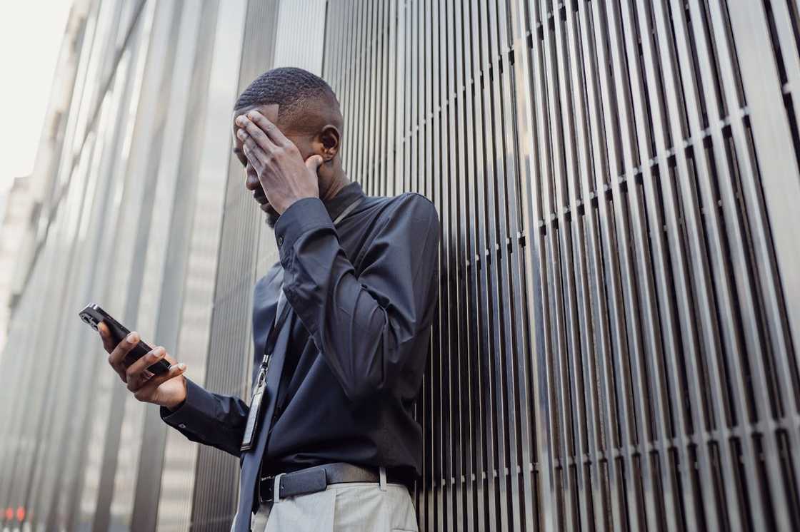 A man stands against a metal wall holding his phone and covering his face.