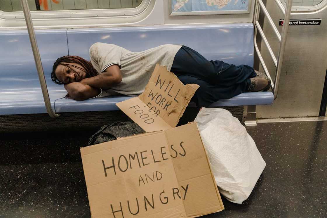 A homeless person is sleeping on a train bench with some cardboard signs. A homeless person is sleeping on a train bench with some cardboard signs.