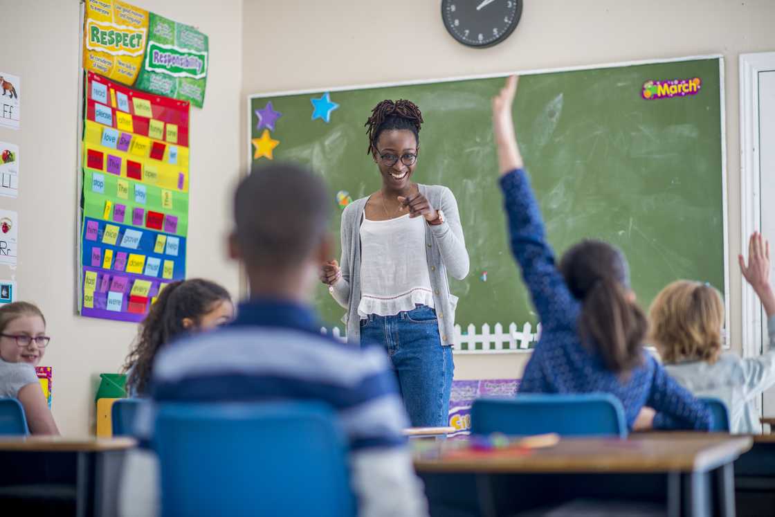 Teacher smiles and points toward students with raised hands in a colourful classroom. Teacher smiles and points toward students with raised hands in a colourful classroom.