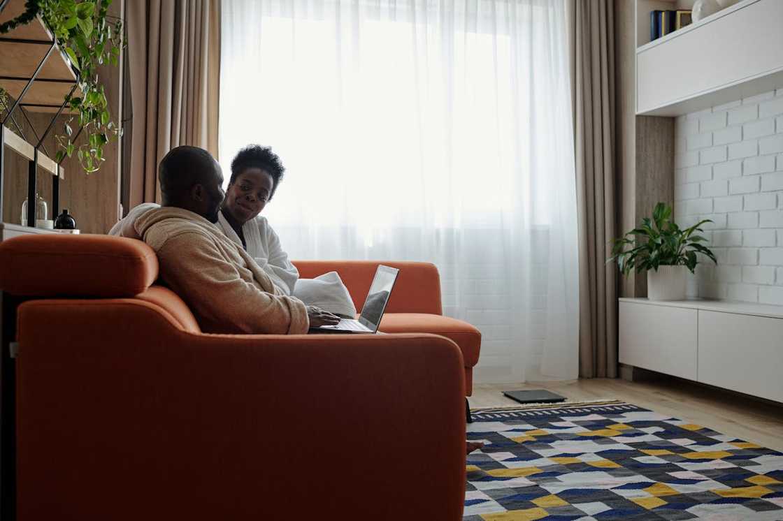 A couple sits together on an orange sofa, talking while one uses a laptop.