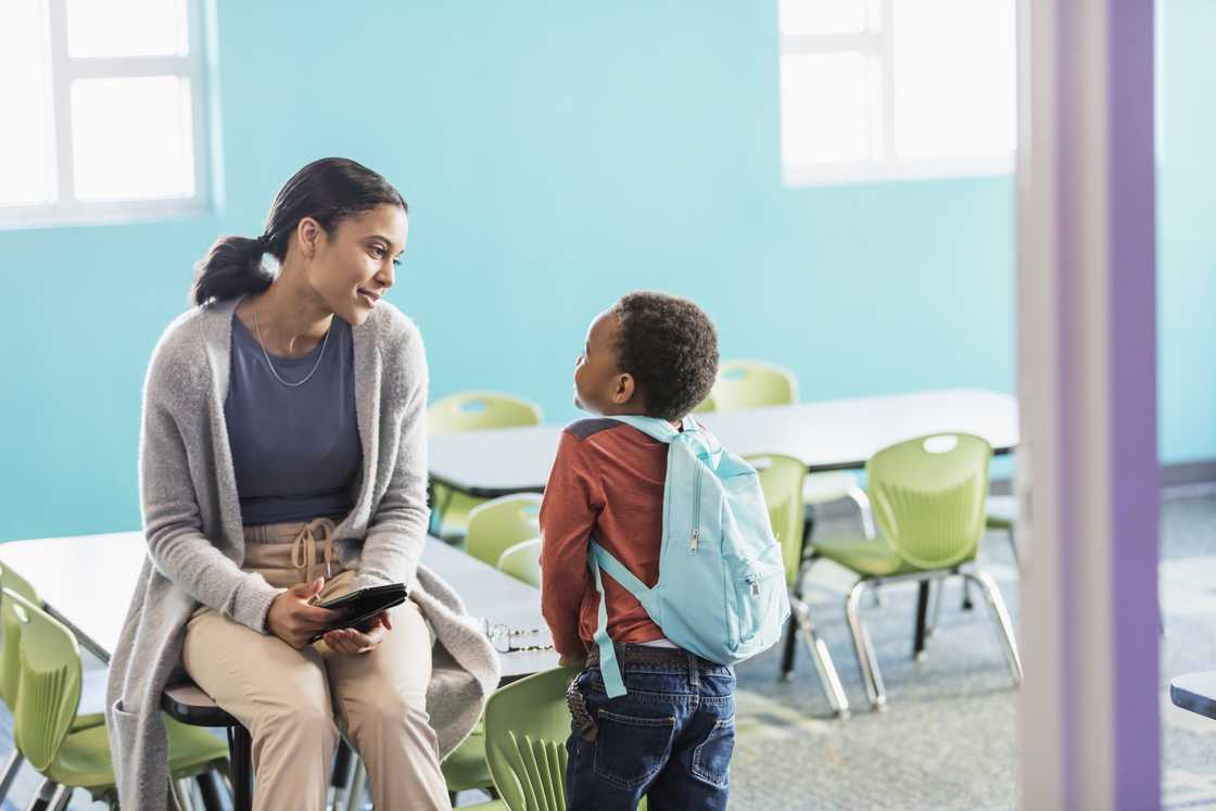 Woman sits on a desk smiling at a young child with a light blue backpack. Woman sits on a desk smiling at a young child with a light blue backpack.