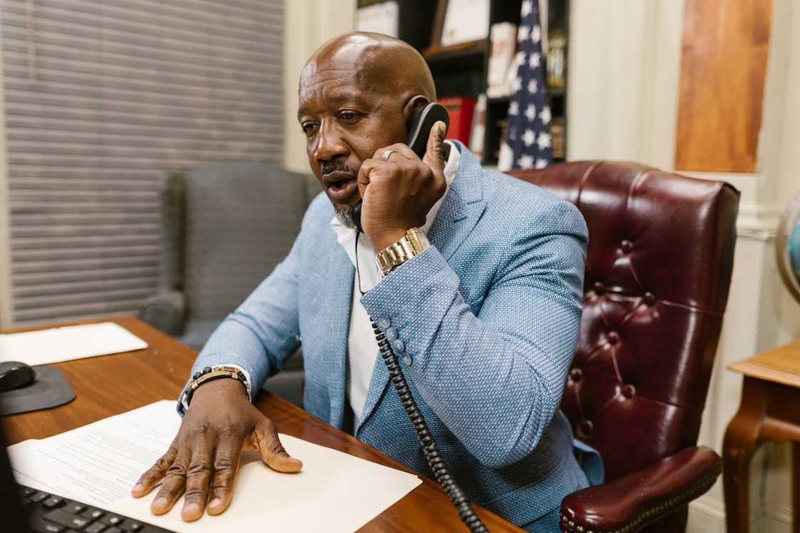 A man in a blue blazer sits at a desk, speaking seriously on a landline phone in an office. A man in a blue blazer sits at a desk, speaking seriously on a landline phone in an office.