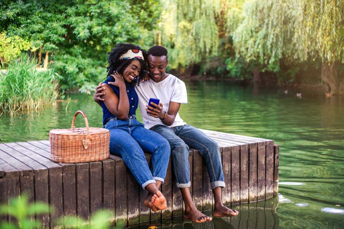 A young black couple sitting on the lake's pier with a picnic basket