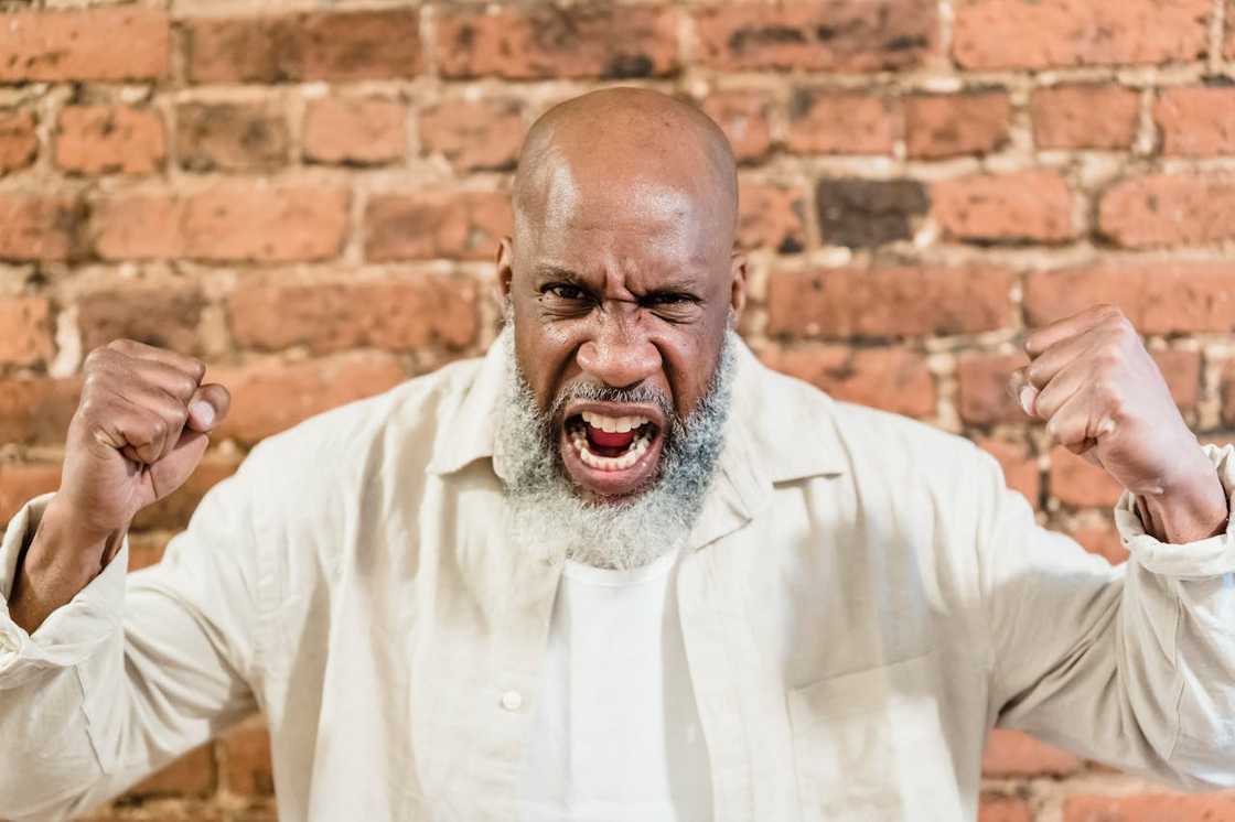 An angry man shouting with fists raised in front of a brick wall.