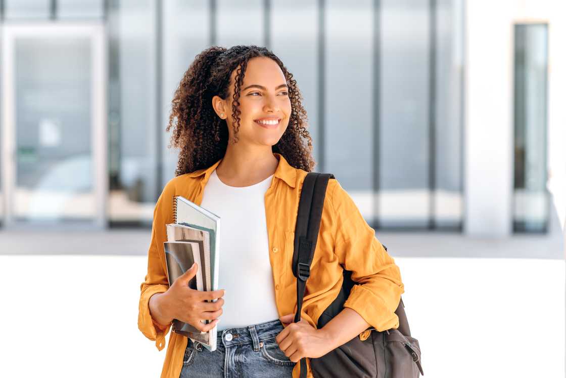 A cheerful female student holding books and notebooks stands near the university campus, looking away and smiling