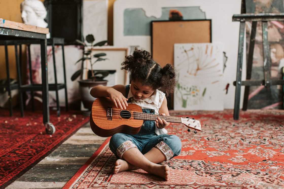 A young girl in a denim jumper sitting on the floor while playing a ukulele instrument A young girl in a denim jumper sitting on the floor while playing a ukulele instrument