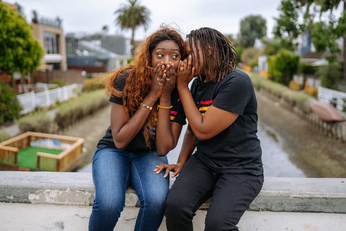 Two women whispering to each other while seated outdoors.