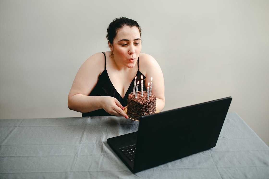 A woman holds a chocolate cake in front of a laptop.