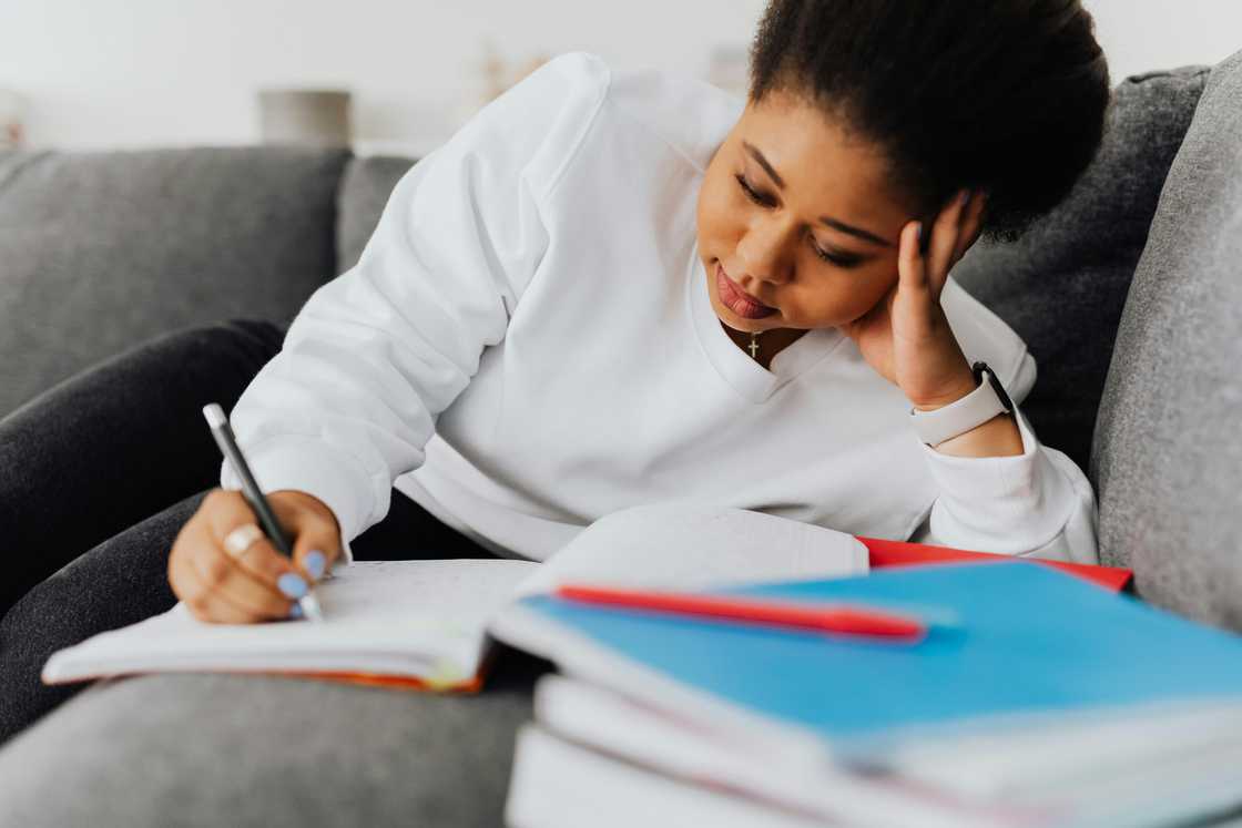 A young woman in a white top doing homework on a grey couch