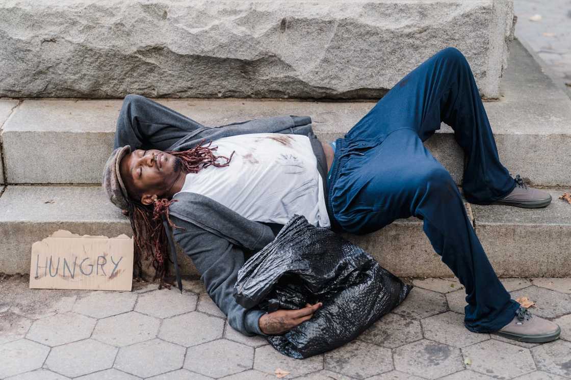 A man lying on stone steps beside a bag and a cardboard sign.