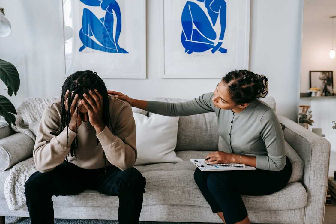 A woman sits beside a man on a couch, gently touching his shoulder as he holds his head. A woman sits beside a man on a couch, gently touching his shoulder as he holds his head.