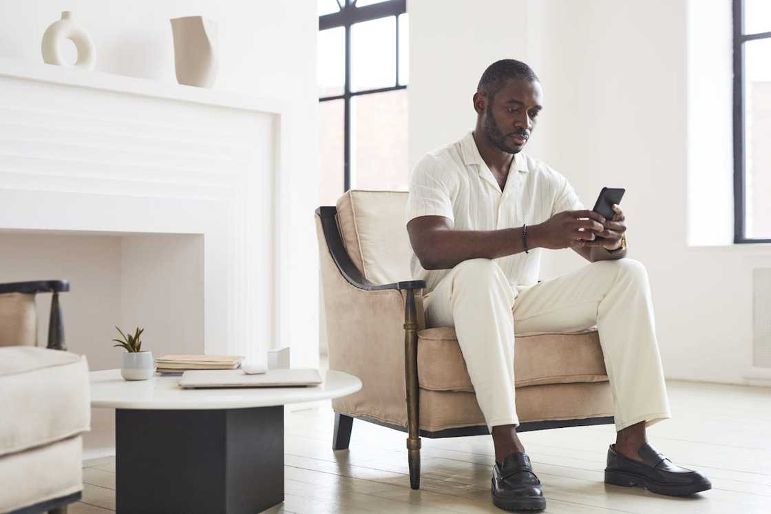 A man sits alone in a chair looking at his phone in a bright living room. A man sits alone in a chair looking at his phone in a bright living room.