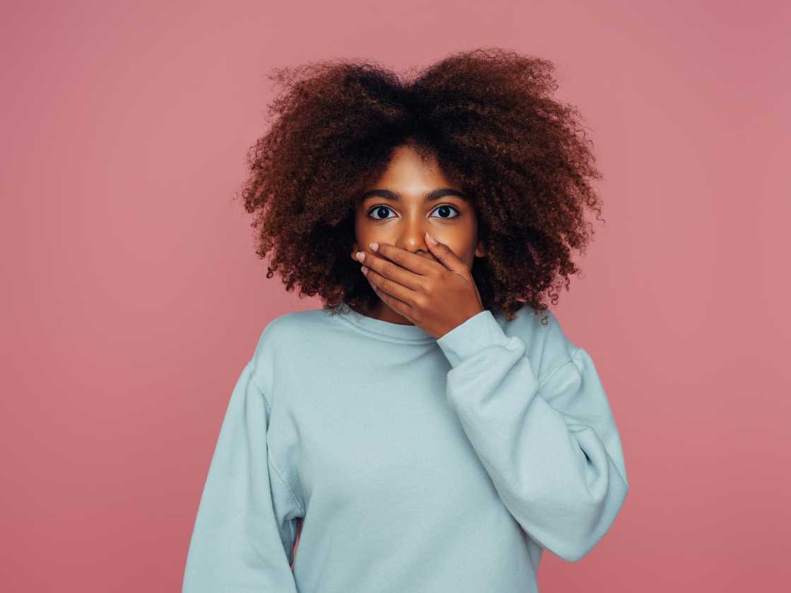 Person in blue sweatshirt reacts with wide eyes and hand over mouth against pink background.