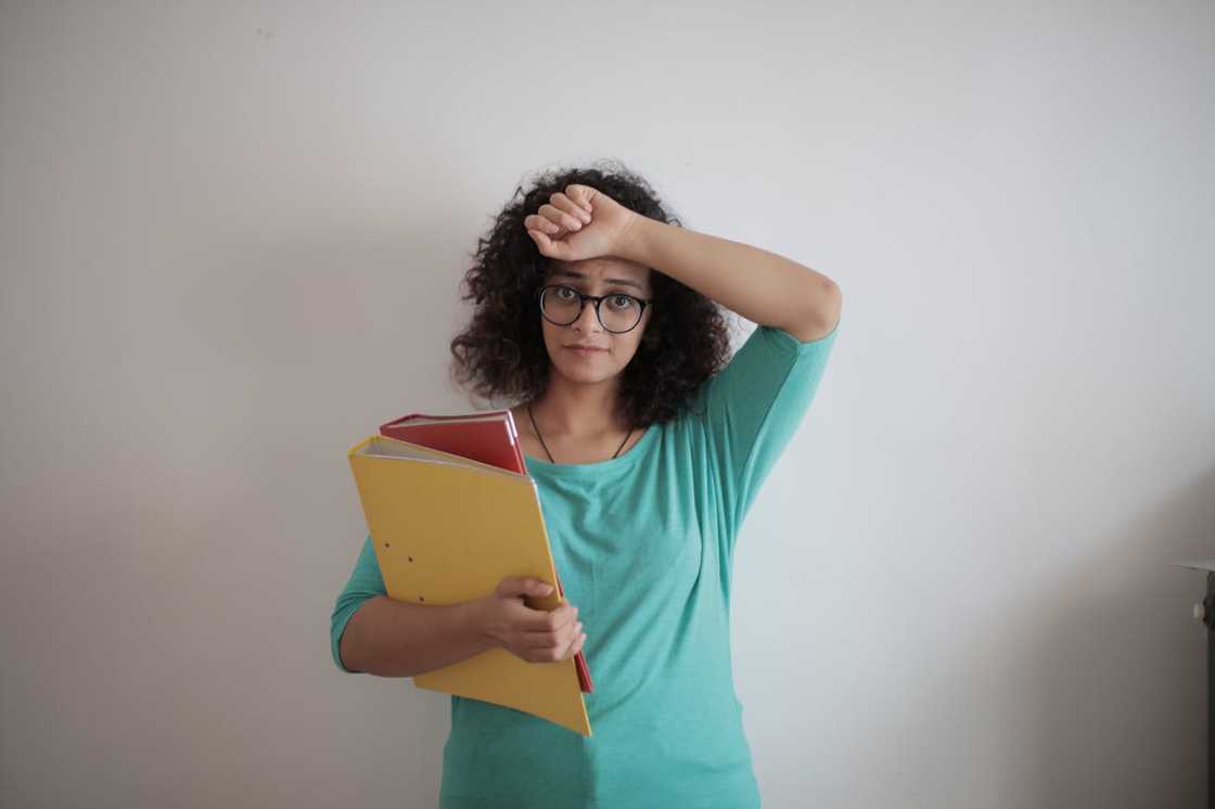 A woman holding folders, raising her arm to her forehead in a stressed gesture. A woman holding folders, raising her arm to her forehead in a stressed gesture.
