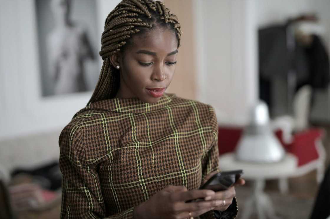 A woman with braided hair looks down at her phone while standing indoors.