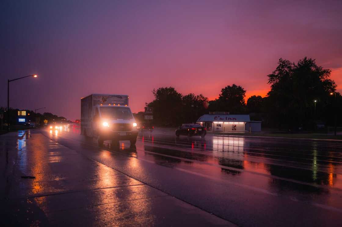 A wet road at dusk shows cars driving under streetlights after rain.