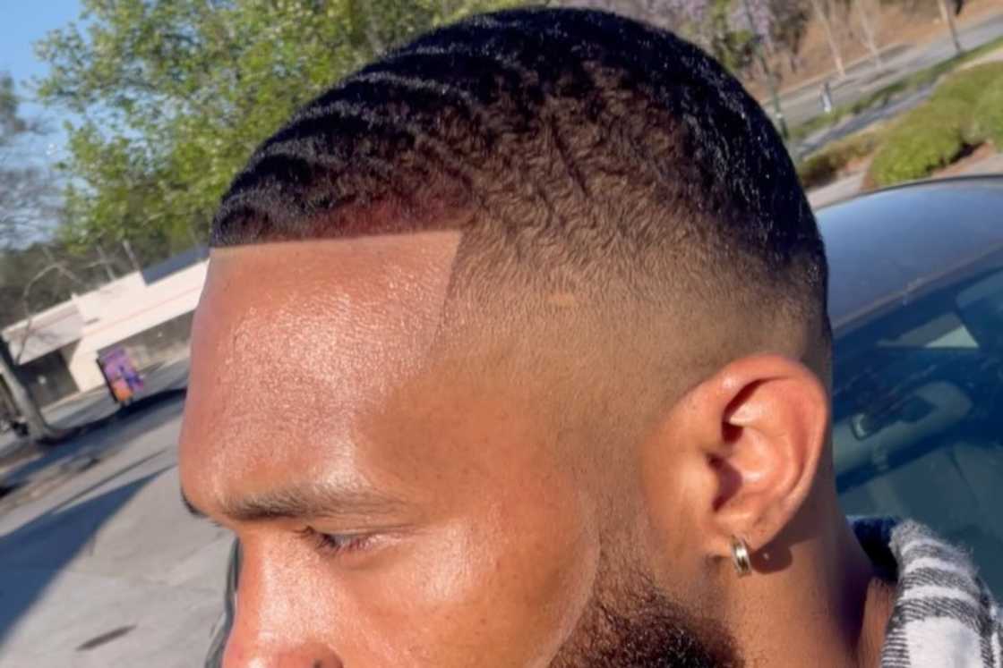 A man with high fade styles is leaning on a car