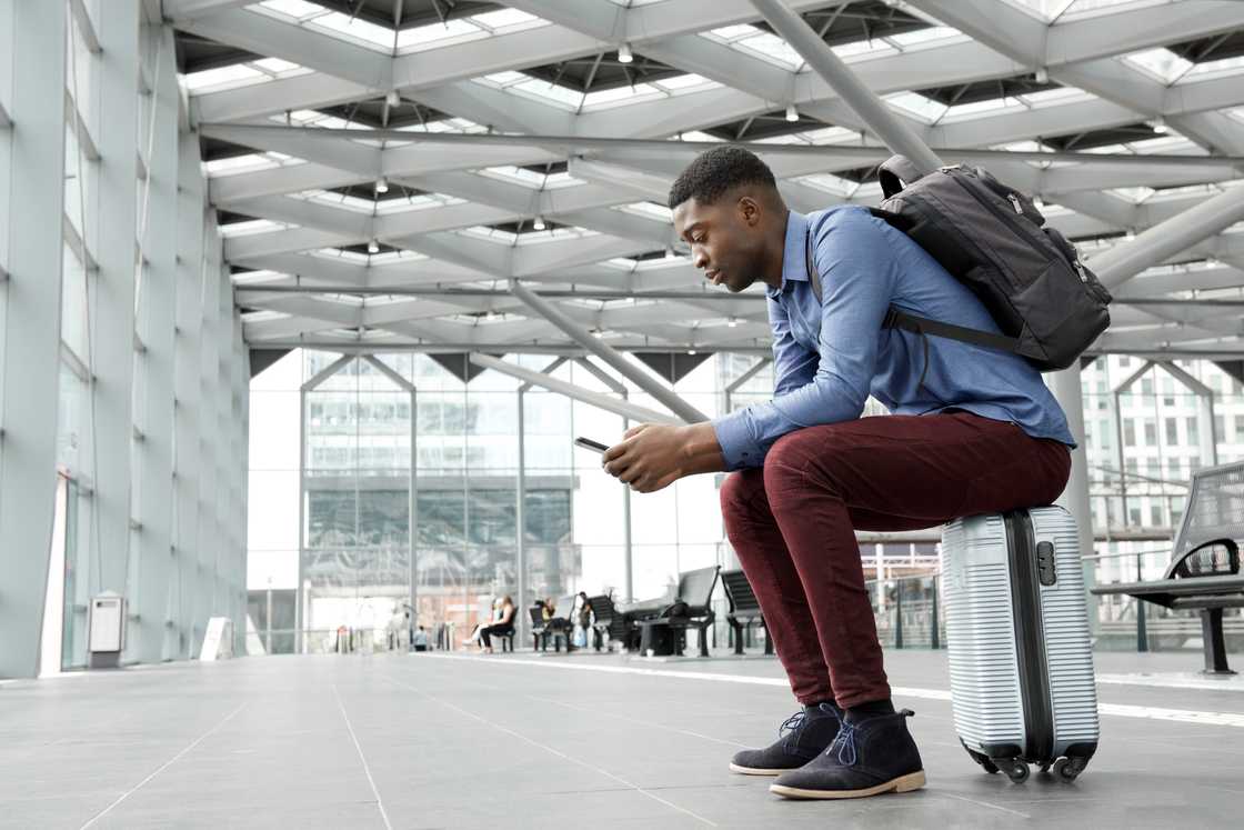 Person sits on a silver suitcase in a modern terminal, looking at a smartphone with a backpack on.