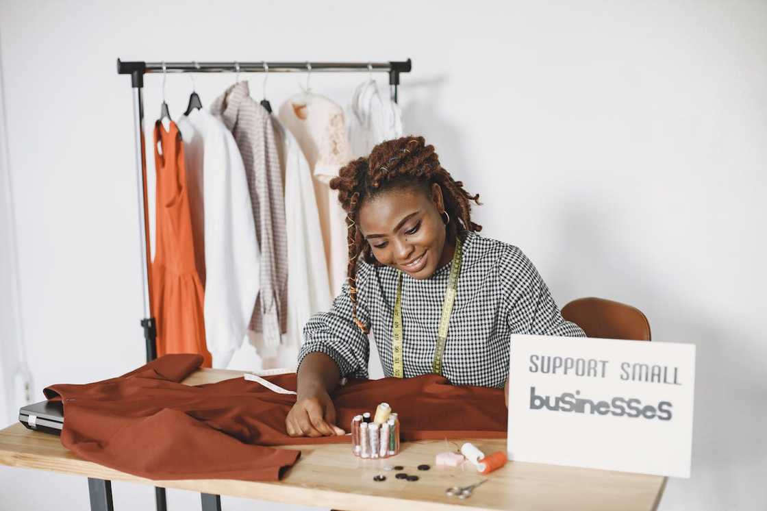A woman sews clothing at a worktable with fabric and tools.