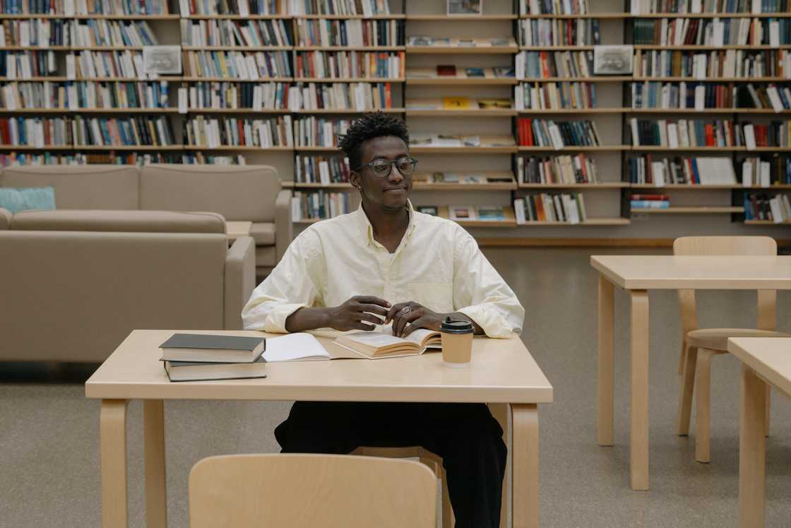 A young man in glasses and a cream shirt is studying at a library