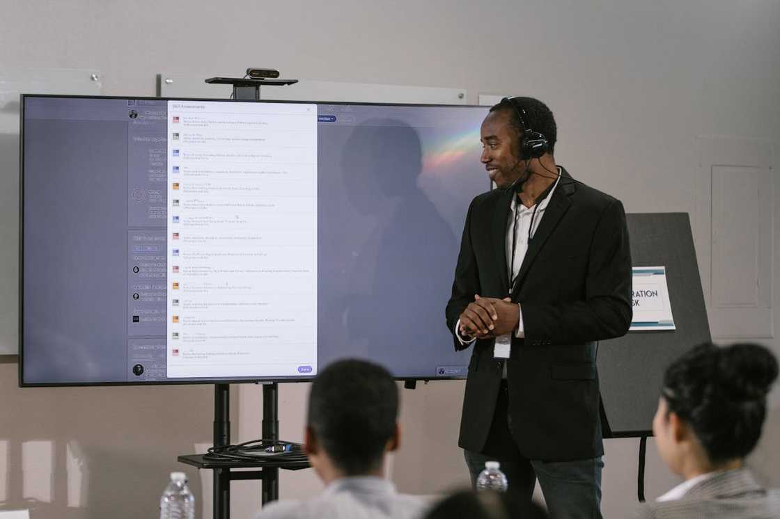 A man wearing a headset presents information on a large screen to a seated audience.