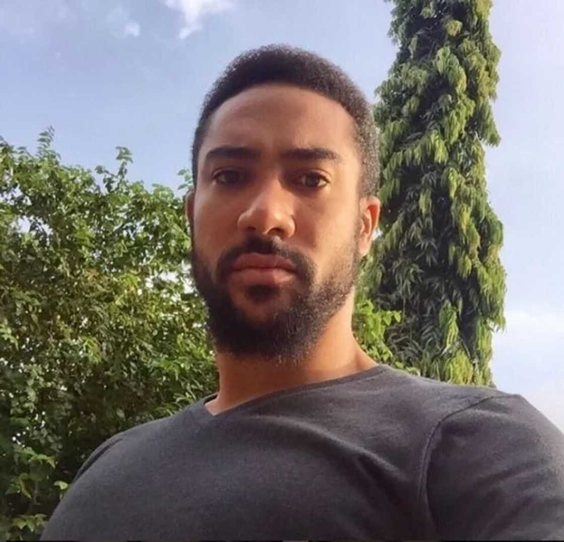 Majid Michel in a grey shirt starring downwards into a camera