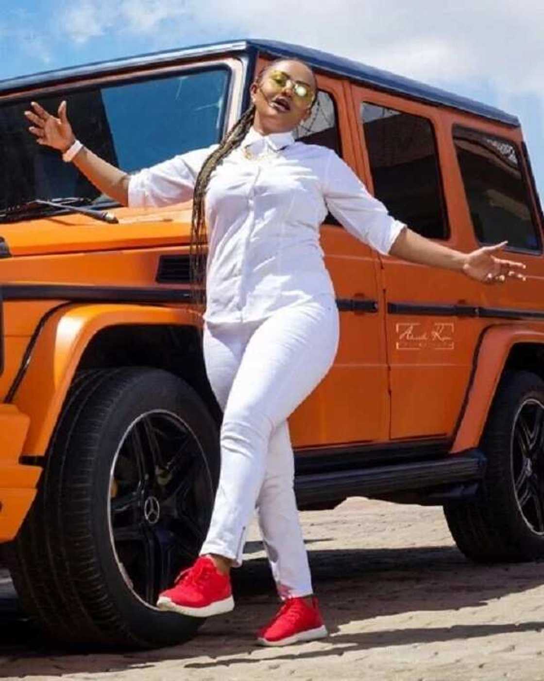 Nana Ama McBrown in a white outfit and red shoes standing in front of an orange jeep