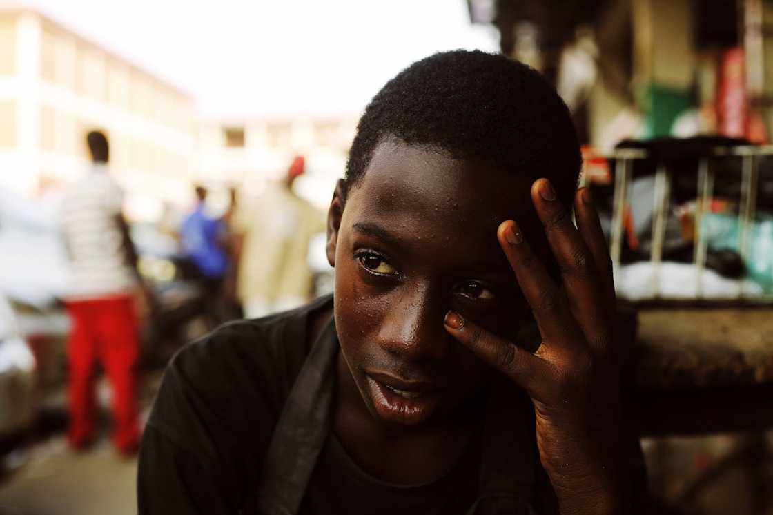 A young boy holding his head, sitting in a crowded outdoor market. A young boy holding his head, sitting in a crowded outdoor market.