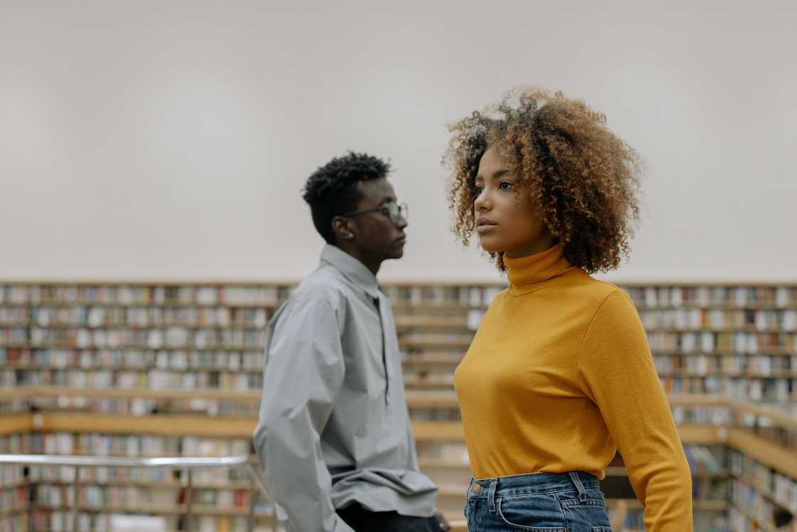 A young man in a blue shirt and a young woman in a mustard top walking past each other in a library