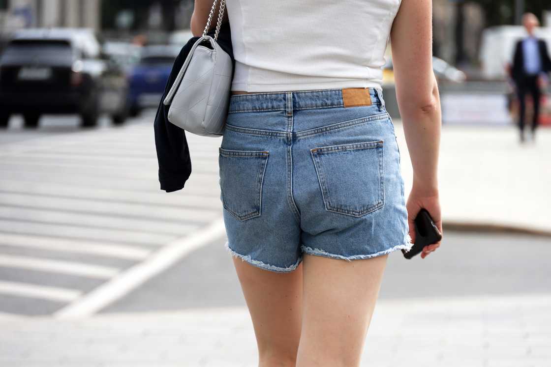 A girl in summer denim shorts walking with a purse and smartphone on a street