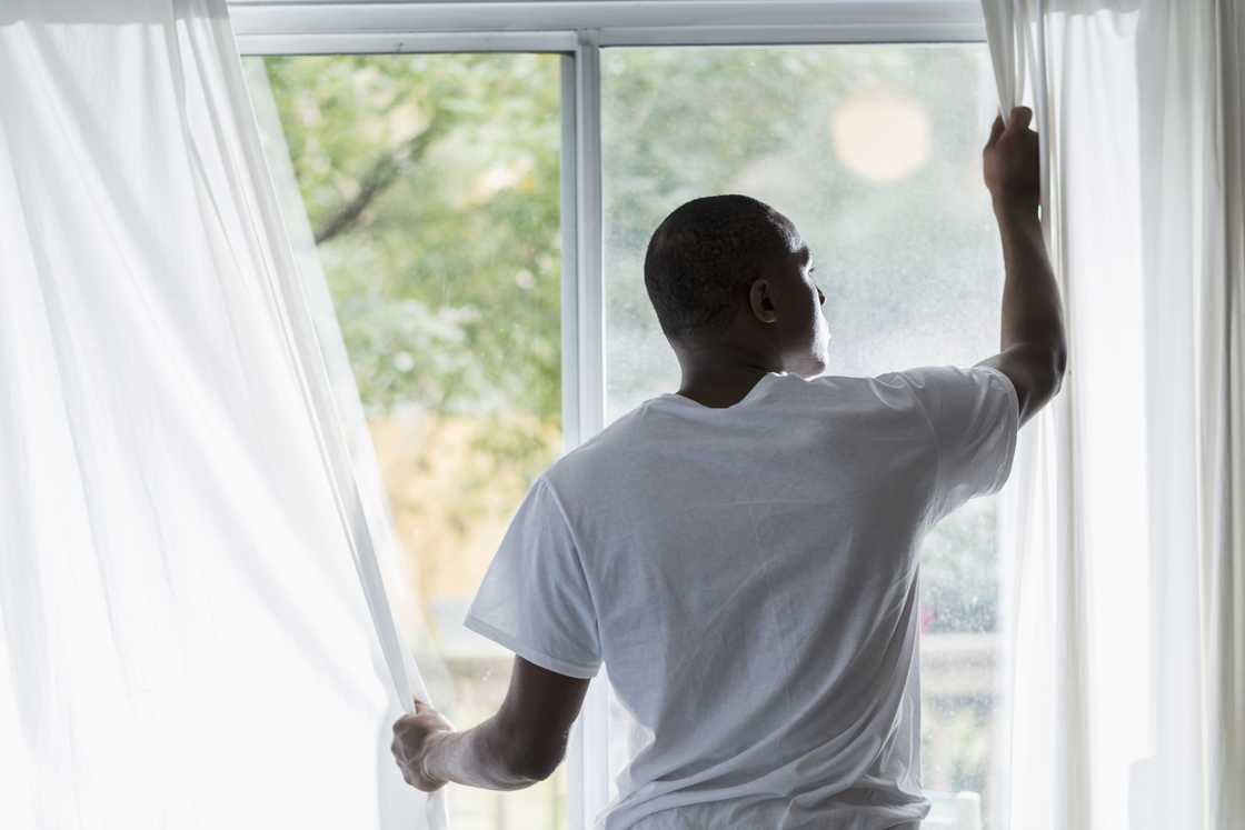 Person opens white curtains, standing in front of a sunlit window with greenery outside.