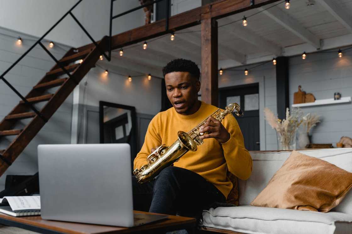 A man holds a saxophone while sitting in front of a laptop.