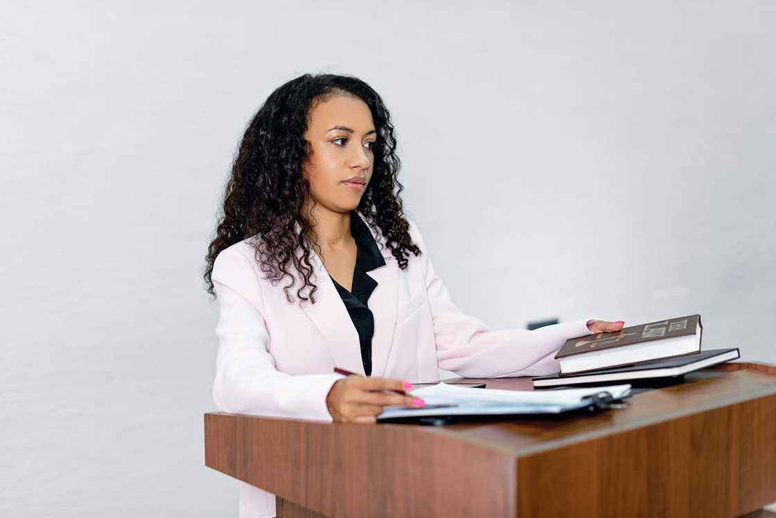 A woman in a pink blazer standing at a podium with books and documents. A woman in a pink blazer standing at a podium with books and documents.