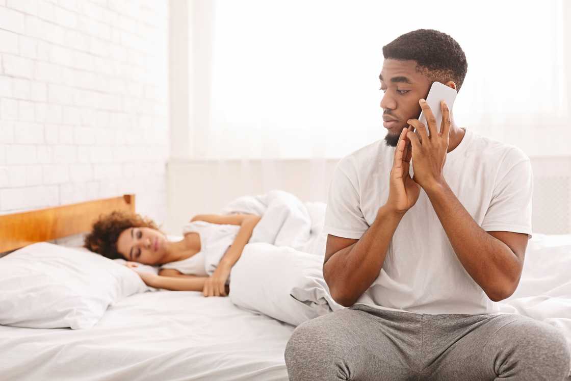 A man on the phone at bedside while partner sleeps.