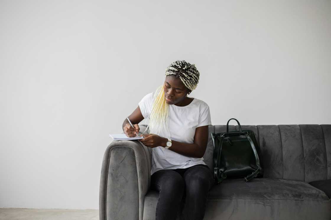 A young lady in braided hair and a white tee is writing on a notebook