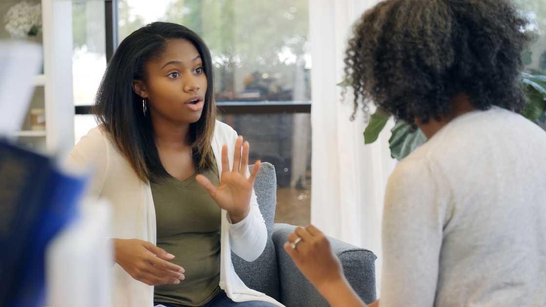 Two people sit in a sunlit room, one gesturing expressively while the other listens attentively.