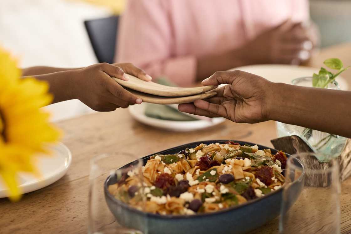 Two people pass stacked wooden plates across a dining table set with pasta salad and a sunflower.
