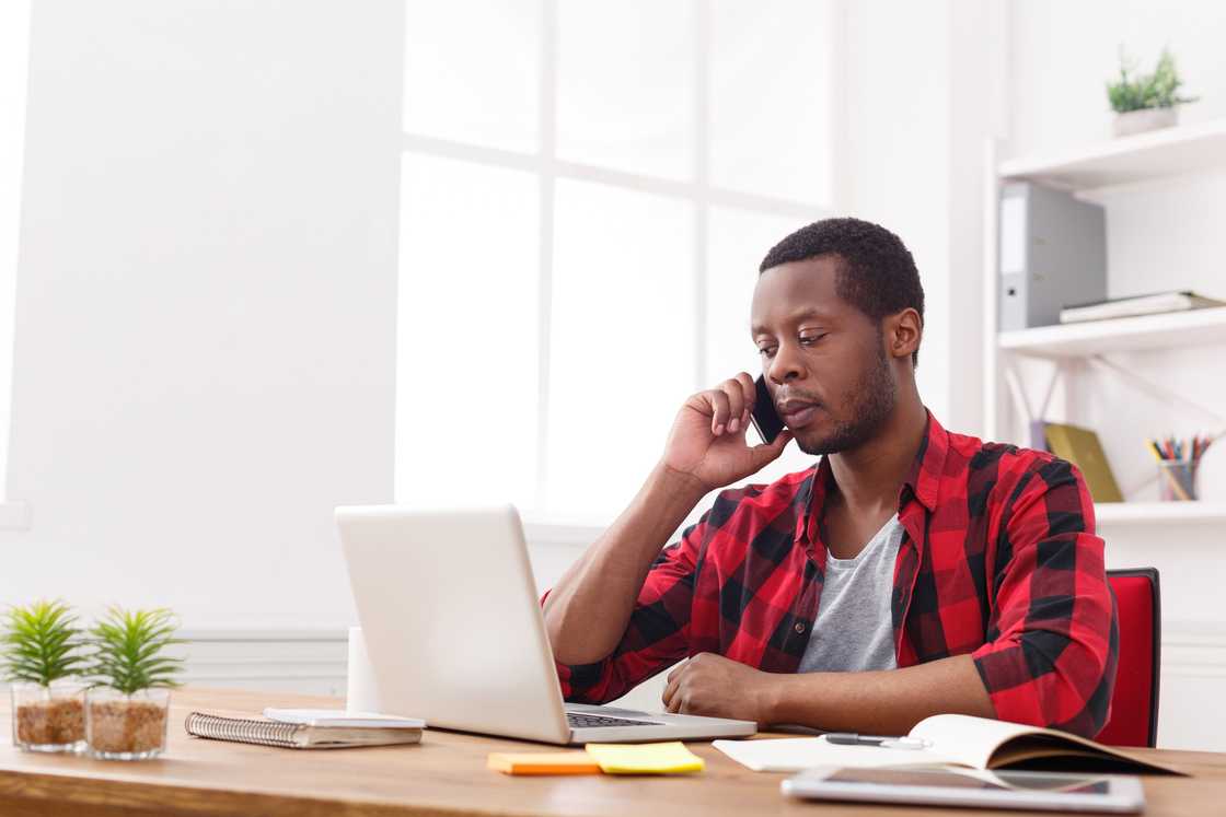 Person in checkered shirt talks on phone while working at a desk with laptop and notes. Person in checkered shirt talks on phone while working at a desk with laptop and notes.