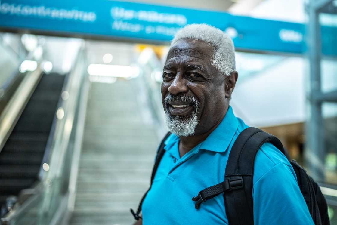 Older person with grey hair and beard smiles while standing in front of an escalator.